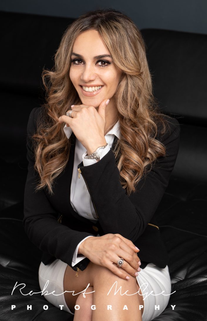 woman is business attire seated on black couch. Chin resting on hand, smiling at camera 019A2878